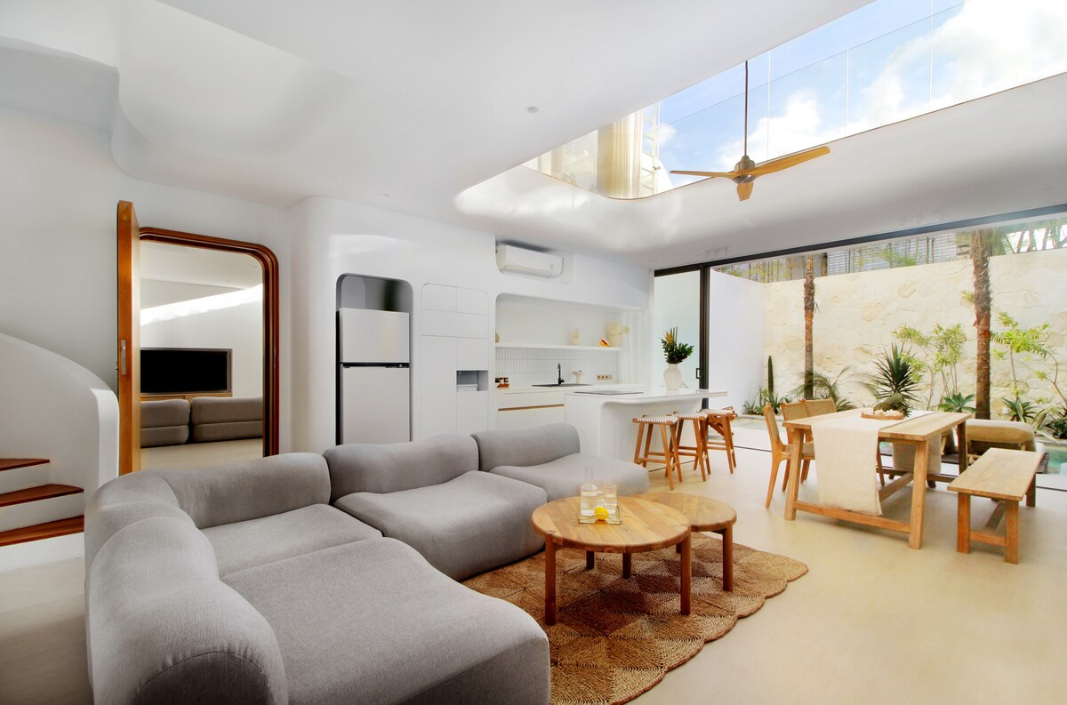 A modern living space features an open layout with a large, sectional grey sofa positioned around a wooden coffee table. A bright kitchen area is visible in the background, complemented by a dining table set with wooden chairs. Natural light flows in through expansive glass doors, revealing greenery outside.