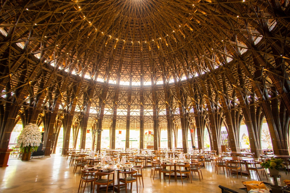 An expansive dining area is enclosed by an intricately designed bamboo structure, featuring a high, rounded ceiling adorned with warm lighting. Round wooden tables are arranged evenly throughout, complemented by elegant table settings, creating a welcoming environment for meals.