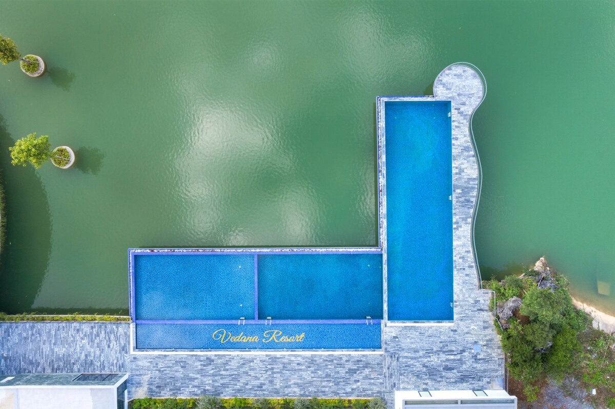 An aerial view of the outdoor swimming pool is presented, featuring a clear blue water surface that contrasts with the green hues of the surrounding lake. The pool is outlined by modern stone decking and blends harmoniously with the natural landscape.