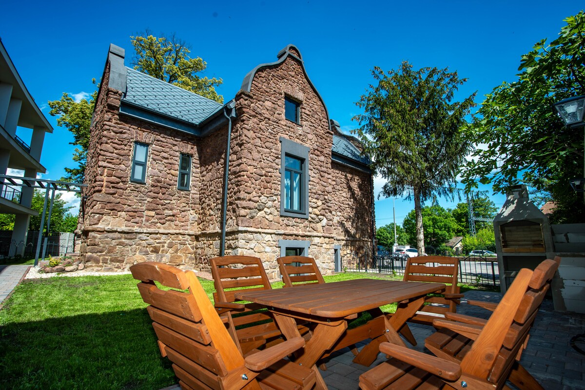 A traditional stone villa is presented with a grassy area in front, featuring a wooden table surrounded by several matching chairs. Lush greenery and trees offer natural shading, while clear blue skies enhance the inviting setting.