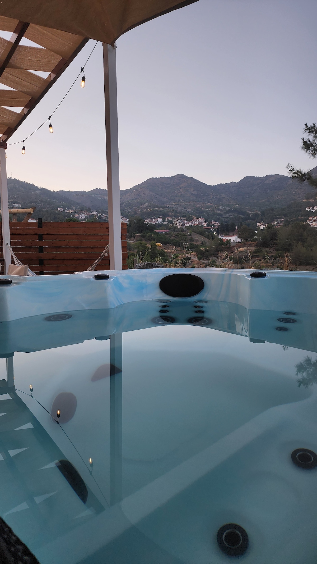 A spacious jacuzzi is featured on a private patio, with clear water reflecting the surrounding mountain landscape. Soft outdoor lighting hangs overhead, enhancing the serene ambiance. The distant mountains are visible in the background, creating a peaceful natural setting.