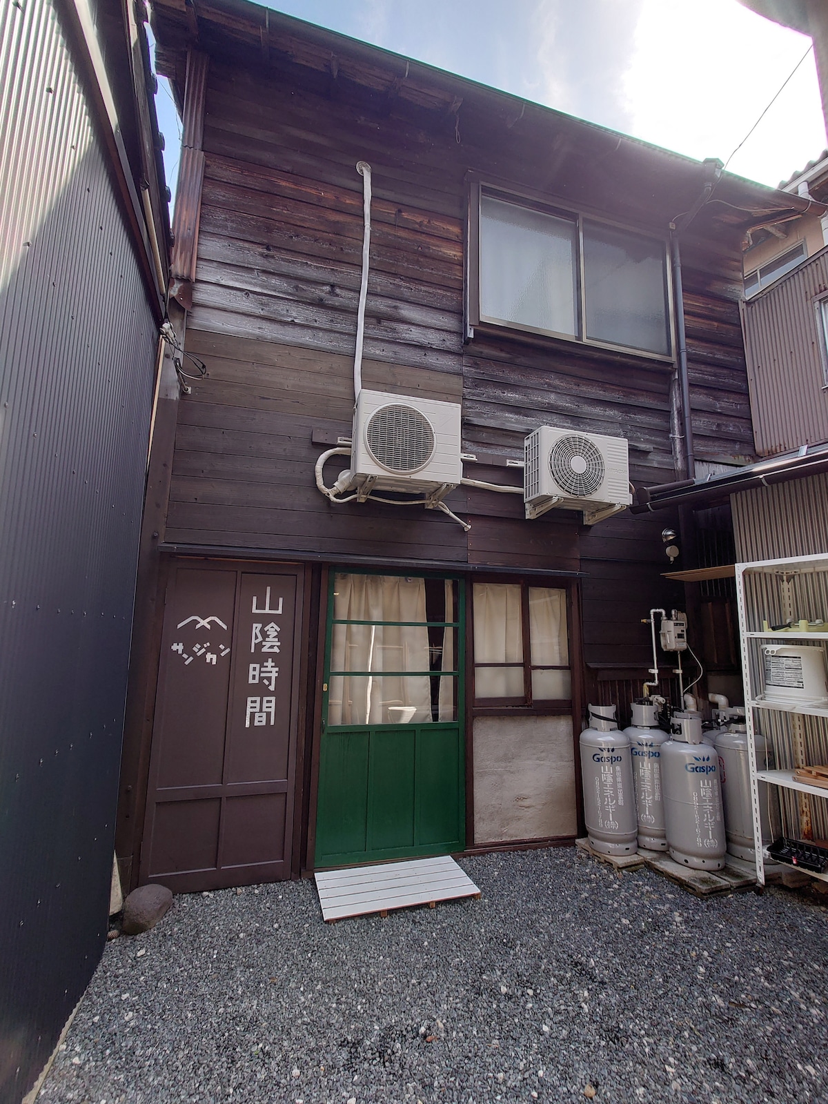 The exterior of the traditional wooden guesthouse features dark wooden panels and a green door entrance. Air conditioning units are mounted above a glass door, with surrounding gravel and some gas cylinders against the wall. A clear sky can be seen above.