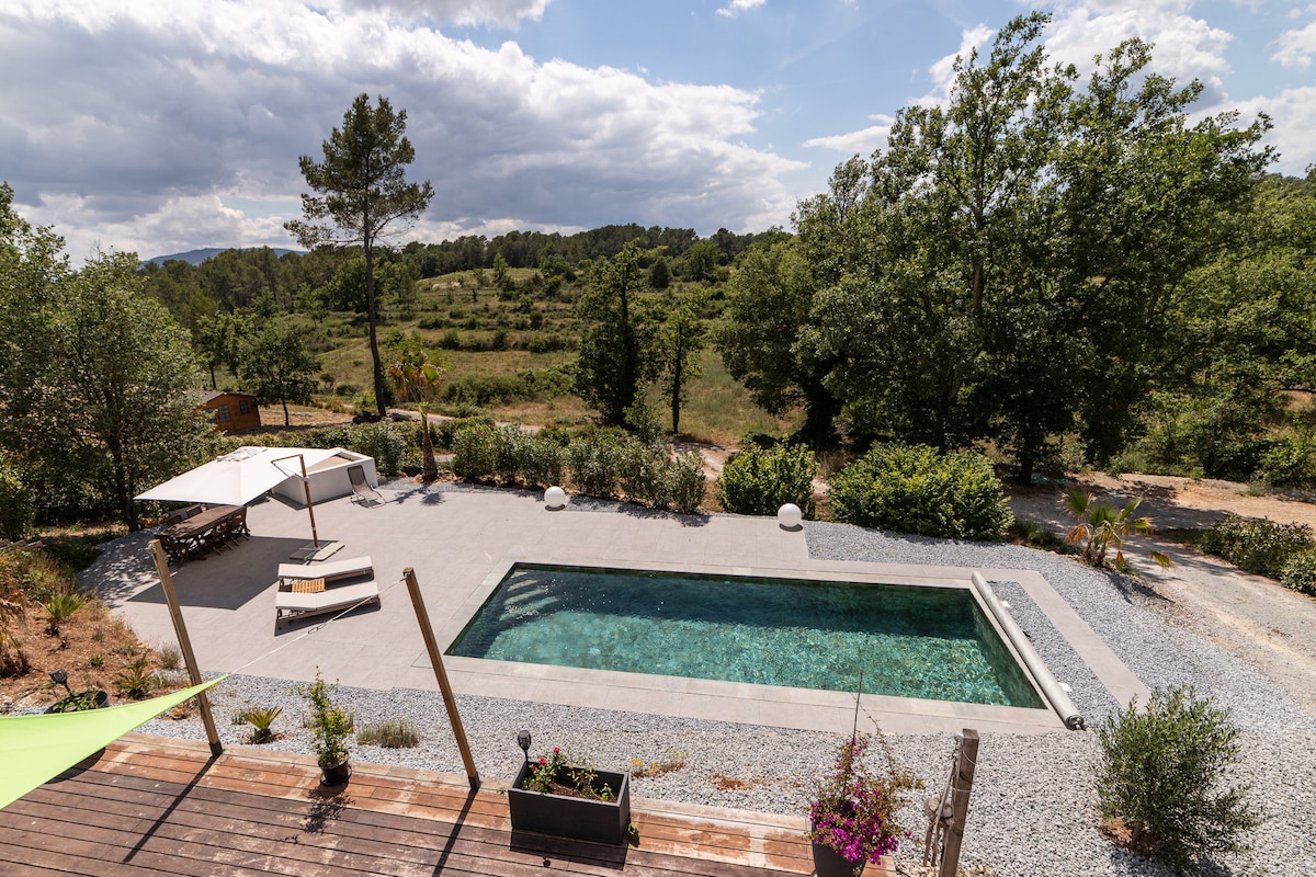 An outdoor view of a rectangular salt-water pool surrounded by a landscaped area. Sun loungers and a shaded cabana offer a place for relaxation. Lush greenery and trees frame the pool, adding to the tranquil setting. The sky features soft clouds, enhancing the serene atmosphere.