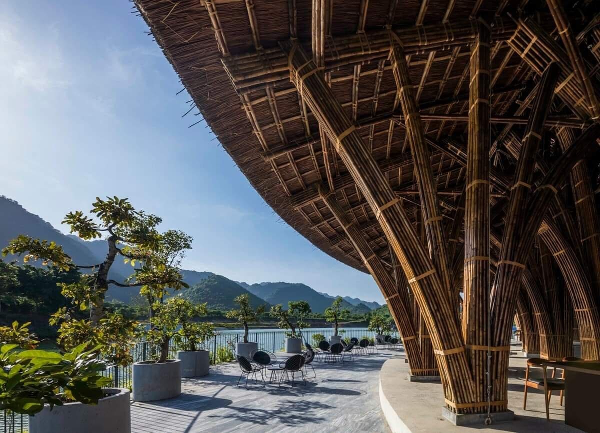 A shaded outdoor seating area features modern chairs arranged on a wide terrace overlooking a calm lake. The building's striking bamboo architecture is visible, with its distinctive roof structure gently curving above. Lush greenery and distant mountains create a serene backdrop.