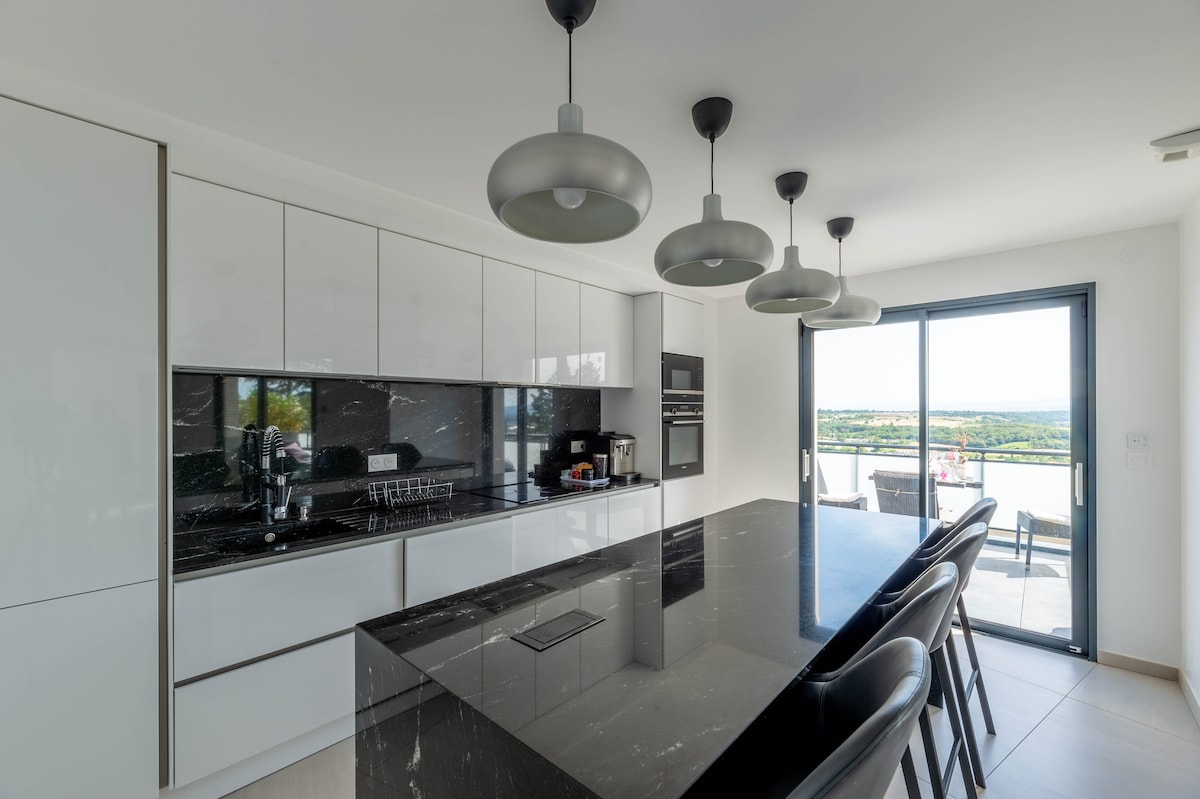 A contemporary kitchen features sleek white cabinetry and a black marble island, equipped with modern appliances. Pendant lights hang above, illuminating the space. Large glass doors provide a view of the outdoor area, creating a seamless connection with nature.