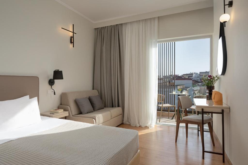 The spacious room features a plush double bed and a cozy sofa bed alongside a small dining table and chairs. Natural light filters in through large windows, highlighting soft, neutral tones. A private balcony is visible, creating an inviting outdoor space.