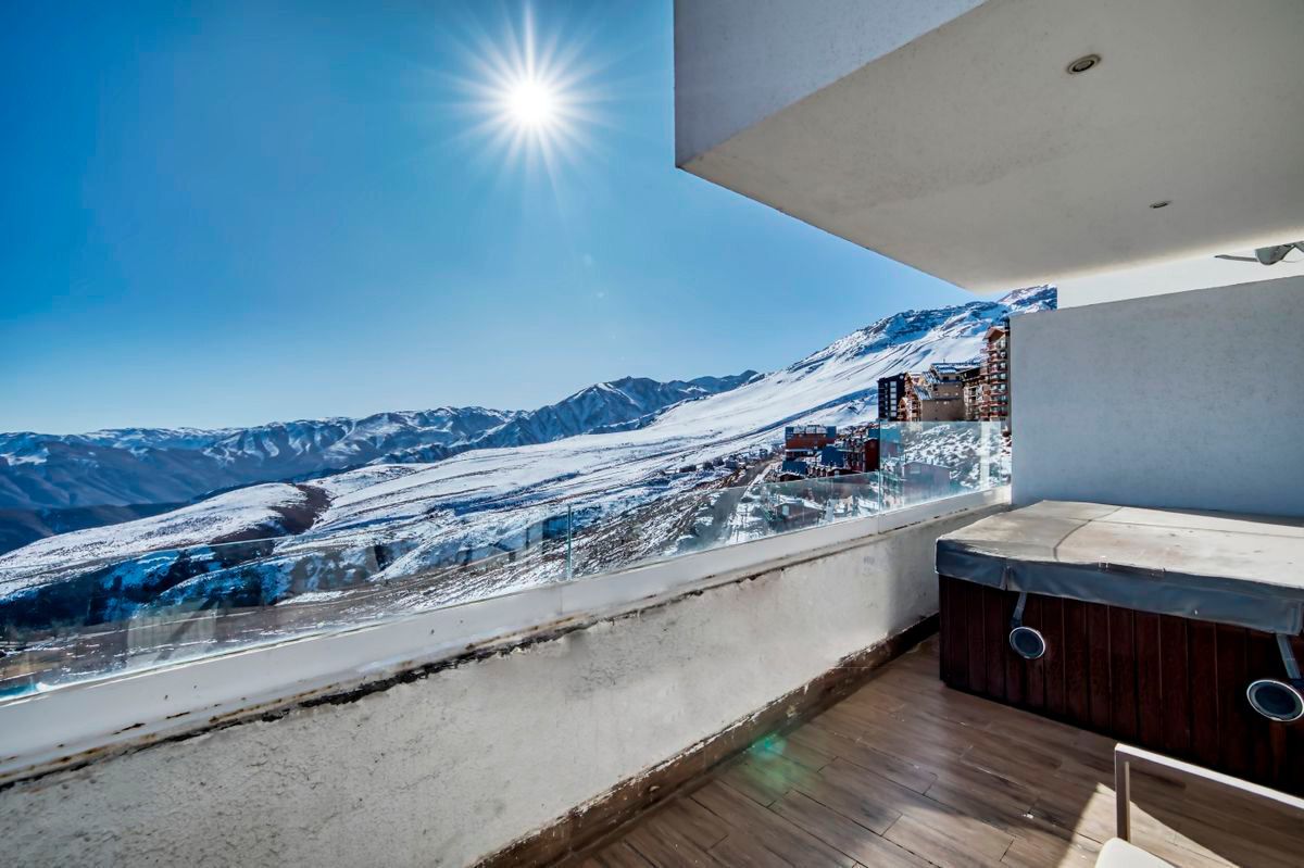 A panoramic view of snow-covered mountains is visible from a spacious balcony. Sunlight reflects off the snow, creating a bright atmosphere. A hot tub is positioned on the wooden deck, inviting relaxation while enjoying the scenic landscape.
