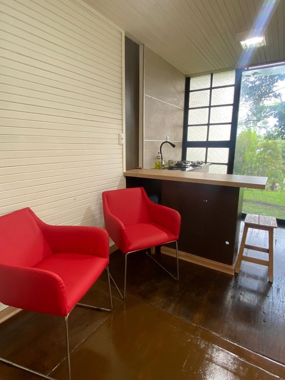 A small kitchenette area features a countertop with a sink and a gas stove, complemented by two bright red chairs. A large window offers views of the surrounding greenery, allowing natural light to brighten the space.