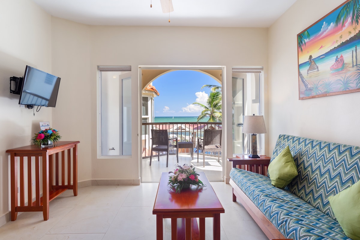 A spacious living area features a patterned sofa and wooden coffee table. Large glass doors open to a balcony with ocean views, where outdoor seating is available. A television is mounted on the wall, and artwork depicting a beach scene adds a colorful touch.