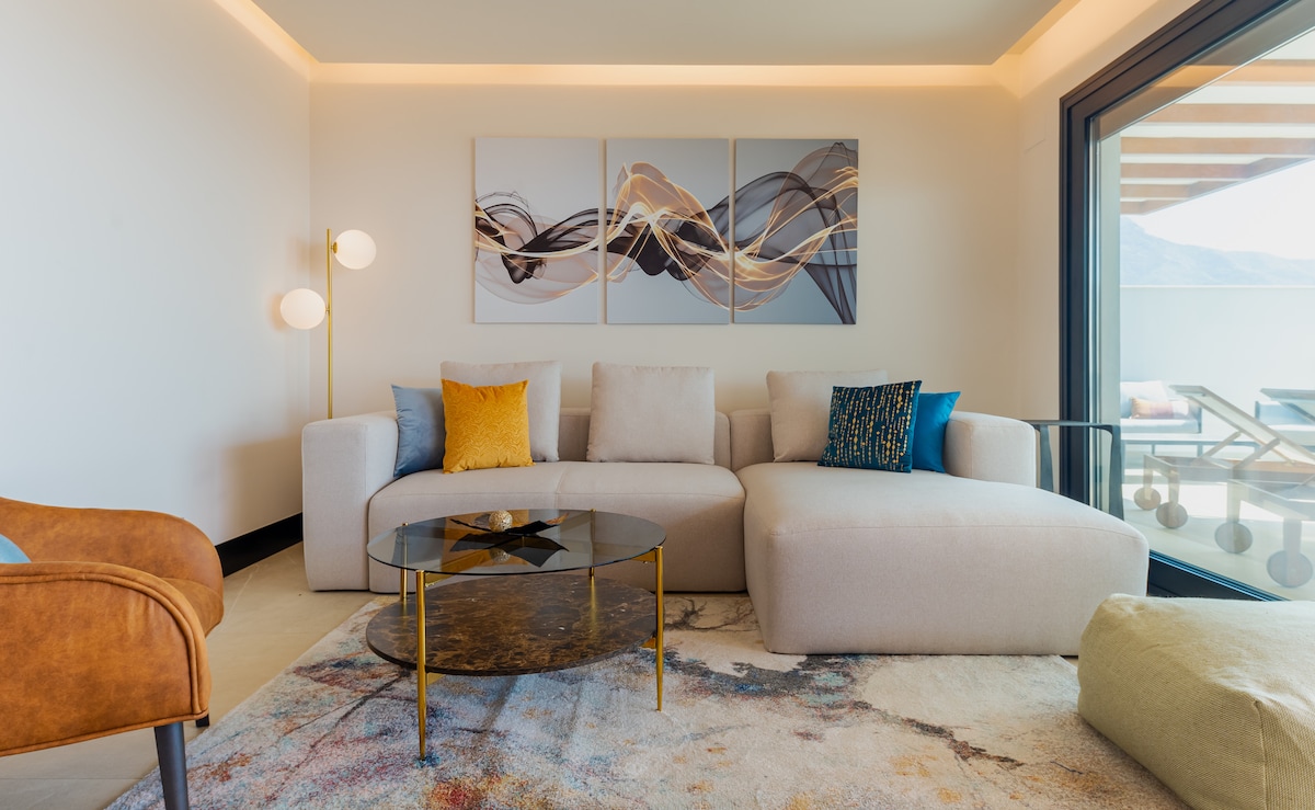 A spacious living area is observed, featuring a modern sectional sofa complemented by accent pillows in various colors. A round coffee table with a dark top sits centrally, while abstract artwork adorns the wall. Natural light filters in through large sliding doors, revealing an outdoor terrace.
