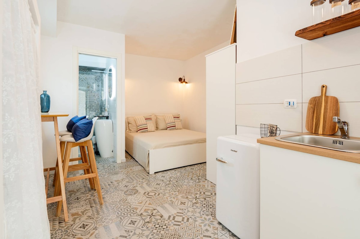 A compact studio features a spacious bed with neutral linens, a small kitchenette with a sink and a mini-fridge, and a dining area with two bar stools. A bathroom door is visible in the background, while patterned tiled flooring adds character to the space.