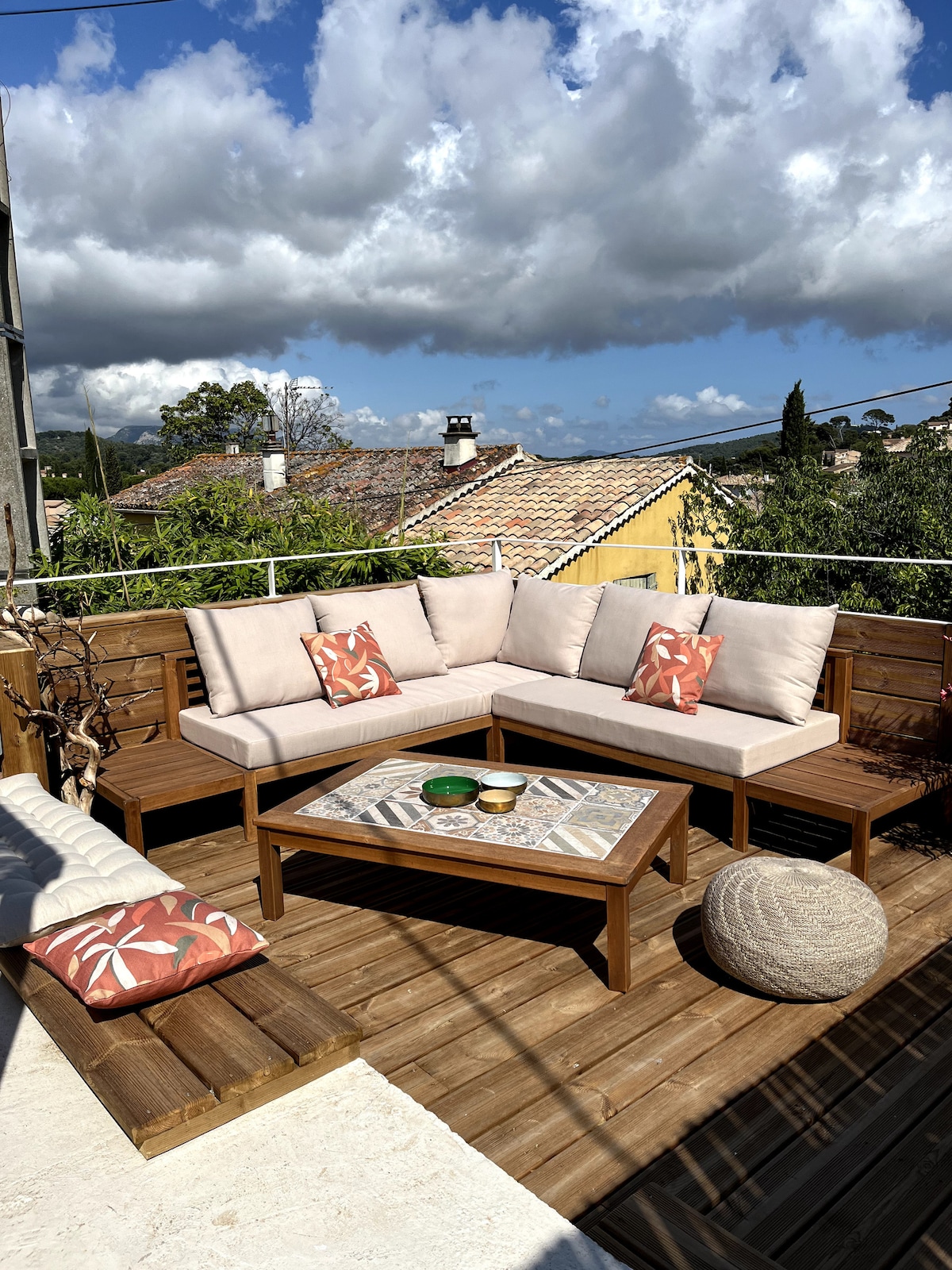 A spacious rooftop terrace is seen, featuring a comfortable outdoor seating area with light-colored cushions and decorative pillows. A wooden coffee table sits at the center, adorned with various decorative items. Lush greenery and rooftops are visible in the background, enhancing the tranquil setting.