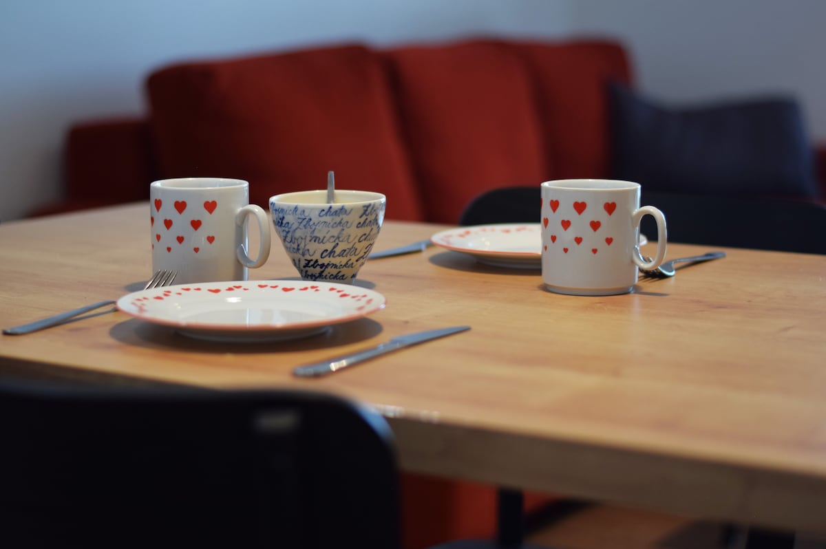 A dining table is set for two, featuring two plates, utensils, and mugs decorated with heart patterns. A bowl sits at the center, surrounded by a warm-colored sofa in the background, creating a cozy dining space.