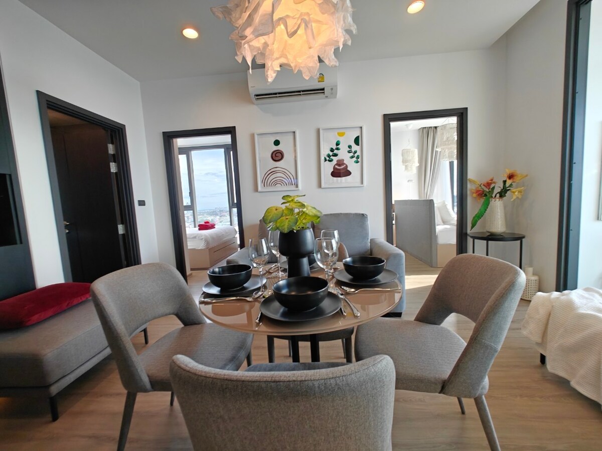 A modern dining area is shown with a round table set for four, featuring elegant dinnerware and glassware. Stylish gray chairs surround the table. Two doors lead to separate bedrooms, while artwork hangs on the wall, adding a pop of color to the neutral decor.