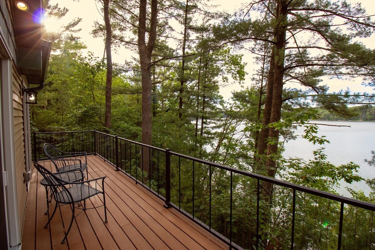A spacious deck overlooks a serene lake, framed by tall pine trees. Two black iron chairs are positioned to enjoy the peaceful views, with sunlight filtering through the foliage, creating a serene and calming atmosphere.