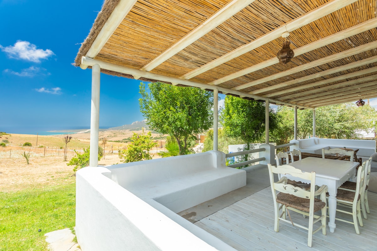 A covered porch features a large dining table surrounded by chairs, providing ample space for gatherings. The area is shaded by a bamboo roof, with views of rolling hills and distant landscapes, creating an inviting setting for outdoor meals.