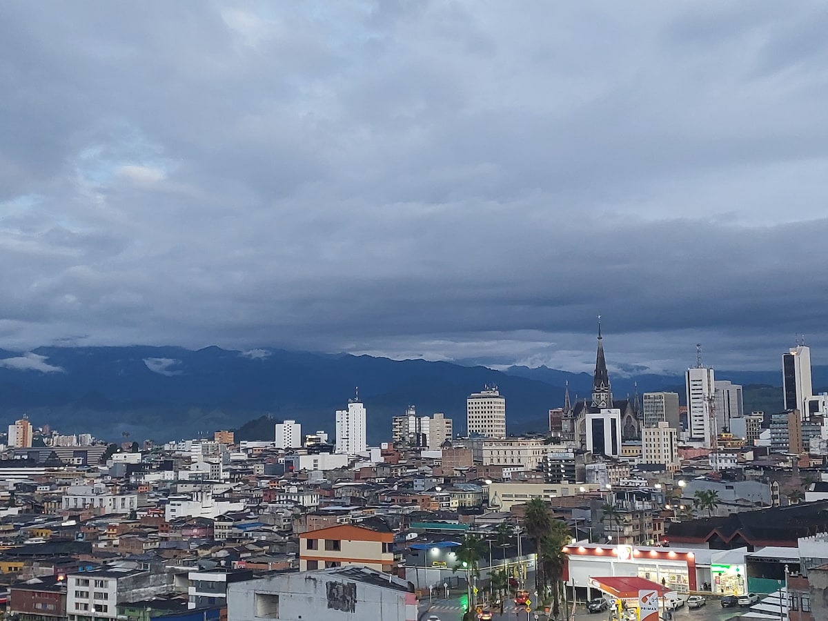A panoramic view of Manizales captures the cityscape under a moody sky. Tall buildings and homes are visible, with the mountainous backdrop providing a dramatic contrast. The image highlights the density of the area, along with the city’s architectural diversity.