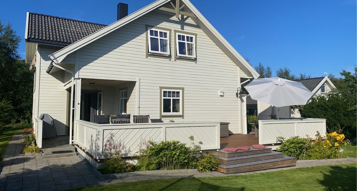 A two-story light-colored house is surrounded by a well-maintained lawn and garden. A spacious deck features comfortable seating and a large umbrella, ideal for enjoying outdoor moments. The house is complemented by large windows that allow ample natural light.
