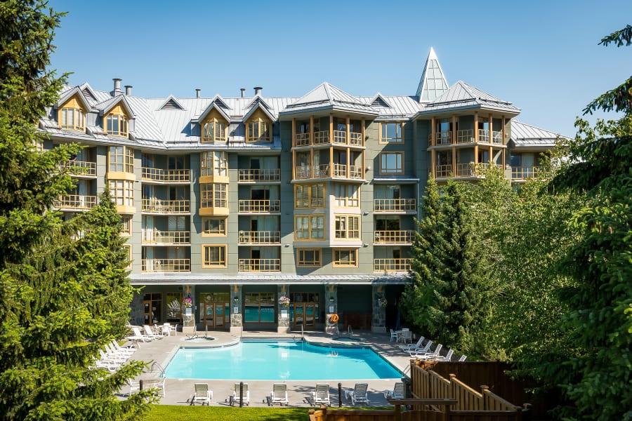 An elegant building with multiple stories showcases large windows and balconies. Surrounding the property, lush greenery provides a natural setting. The outdoor heated pool is centrally located, surrounded by lounge chairs, inviting relaxation under a clear blue sky.