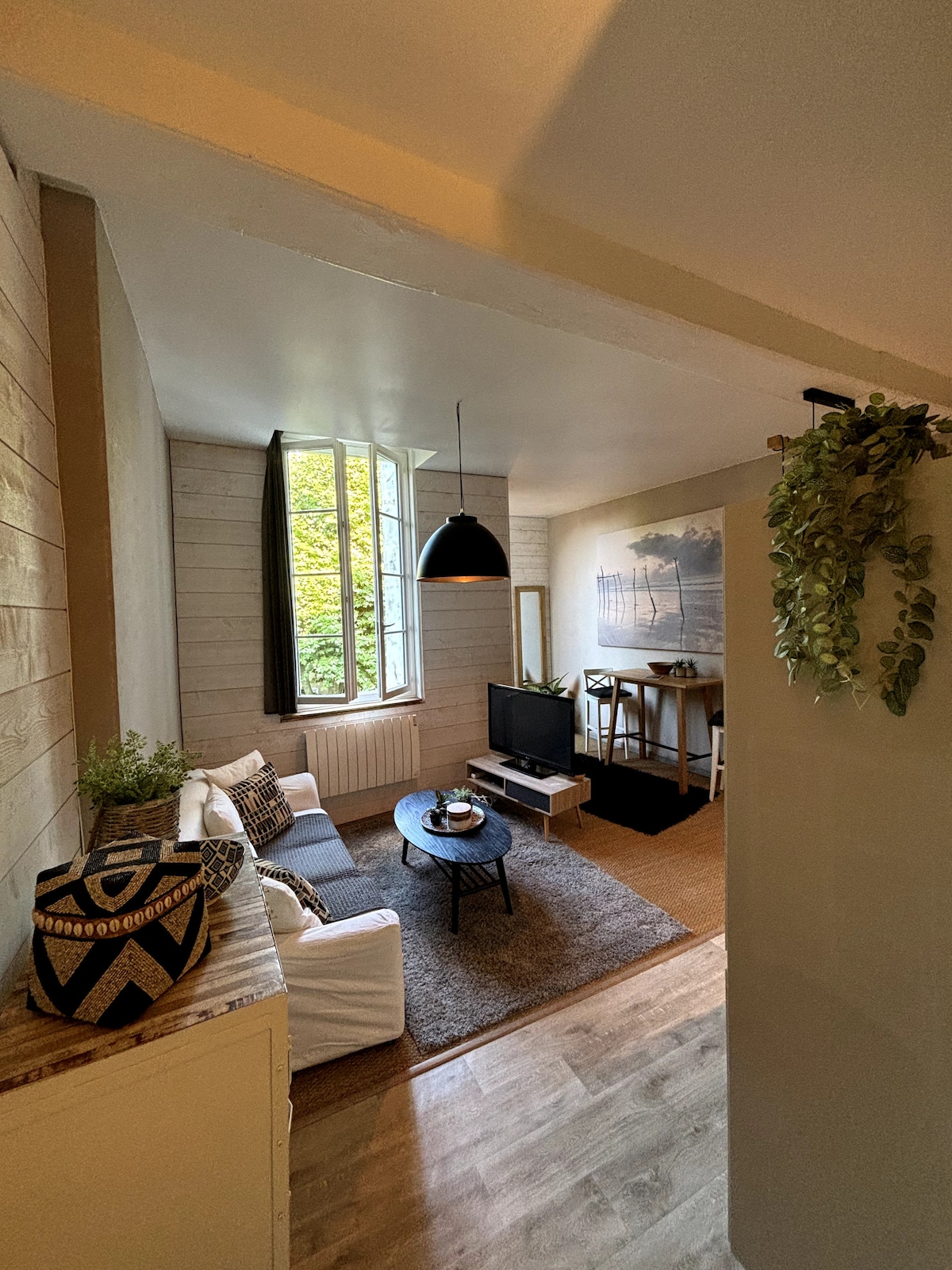 A living room space is shown with a comfortable seating area featuring a white sofa and a dark coffee table. Natural light enters through a large window, which offers a view of greenery outside. A flat-screen television and minimalistic decor enhance the room's cozy atmosphere.