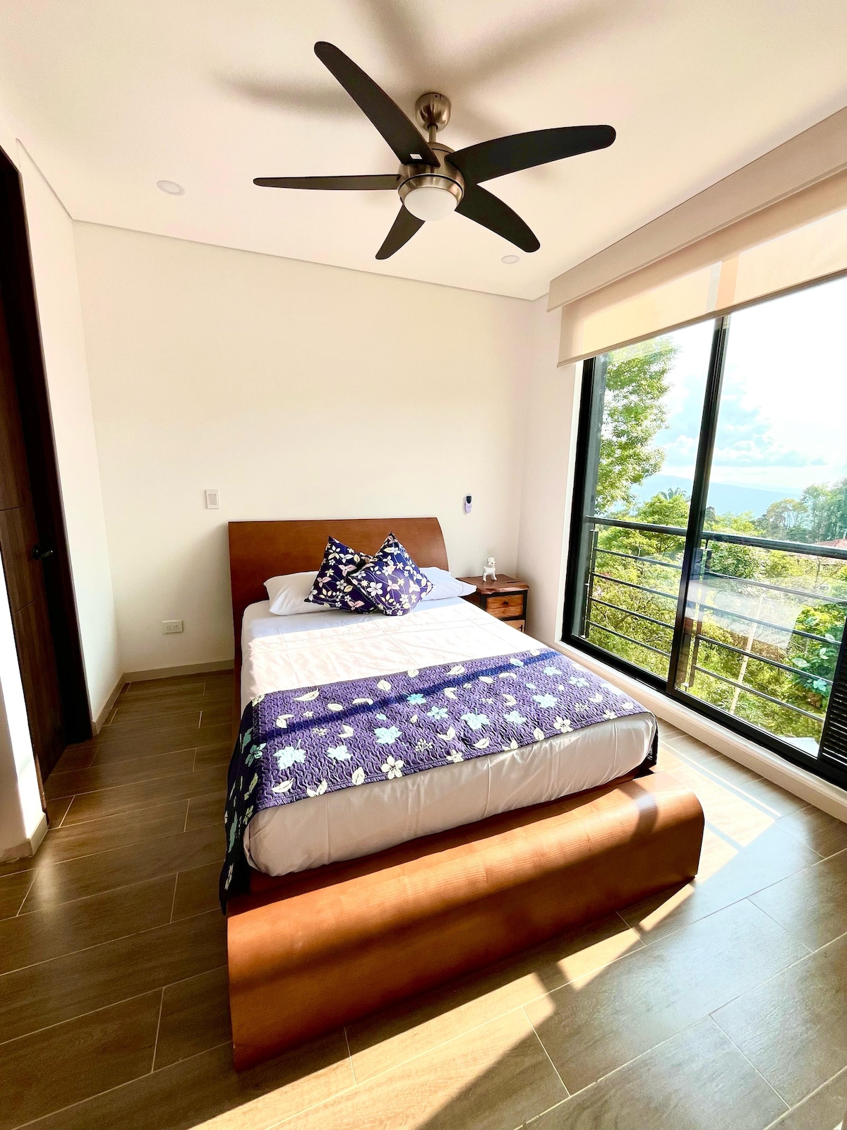 A well-lit sleeping area features a wooden bed with a plush mattress and decorative pillows. A side table holds a lamp, while large windows offer outdoor views and natural light. A ceiling fan facilitates airflow in the space.