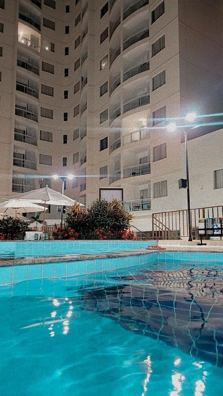 The image showcases a sparkling pool surrounded by vibrant landscaping, with the towering building illuminated by soft lighting above. Leisure areas are visible, featuring umbrellas and lounge chairs, creating a relaxing atmosphere for guests to enjoy.