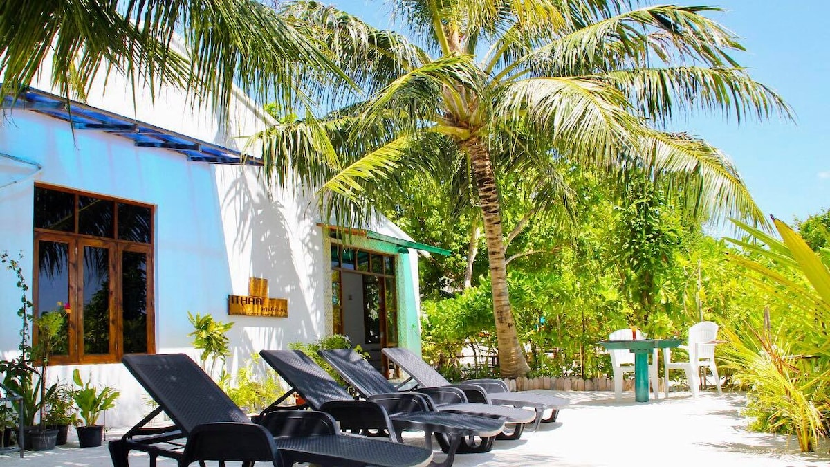 Lounge chairs are arranged under swaying palm trees, offering a space for relaxation. Sunlight filters through vibrant greenery, which enhances the serene outdoor environment. A small table and a chair are positioned nearby, creating a welcoming area for guests to enjoy the surroundings.