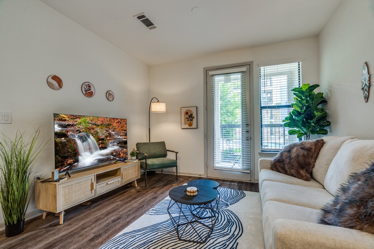 A cozy living area is furnished with a light-colored sofa and an accent chair. A television displays a serene landscape, and a decorative coffee table sits centrally on a patterned rug. Natural light enters through a sliding glass door, which leads to a small outdoor balcony.