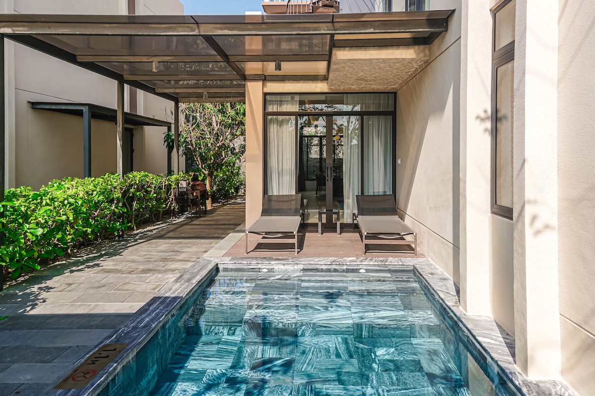 A private outdoor area features a serene pool surrounded by natural stone tiles. Two lounge chairs are positioned beside the pool, with greenery lining the perimeter. Large glass doors provide a view into the interior of the villa, enhancing the connection between outdoor and indoor spaces.