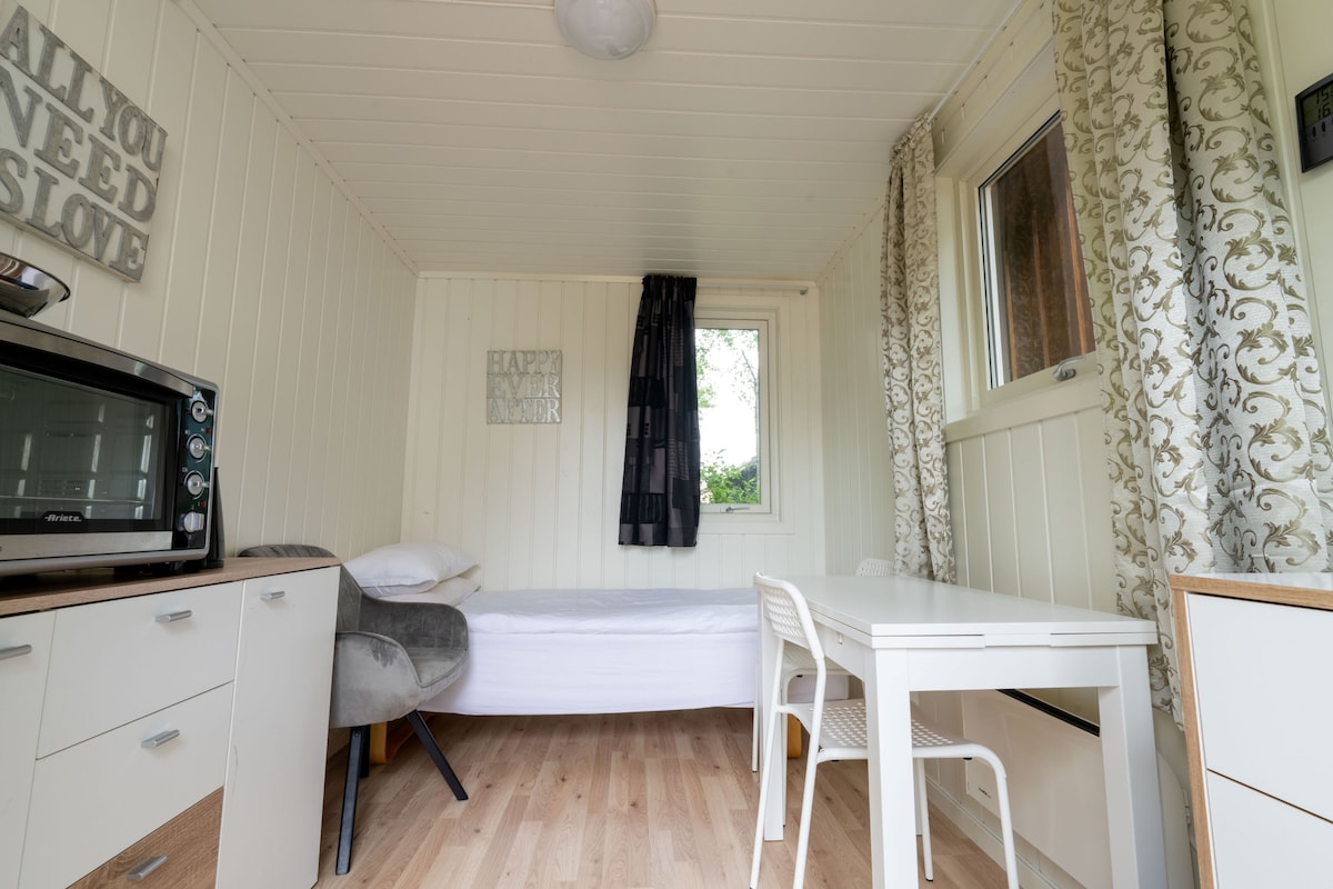 A compact living space is presented, featuring a single bed with fresh linens and a cozy chair. A small white table and two chairs provide a dining area. Light streams through a window adorned with sheer and patterned curtains, enhancing the room's inviting ambiance.