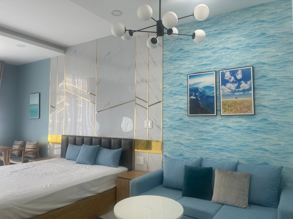 A spacious bedroom is featured with a king-size bed against a decorative wall. A comfortable sofa area includes decorative cushions, while two framed landscape prints complement the design. Natural light illuminates the room, enhancing the calming ocean-inspired color palette.