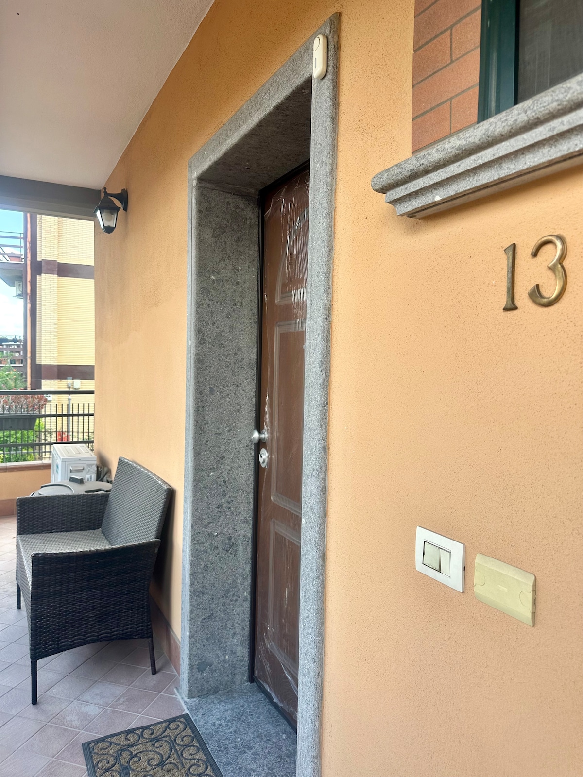 The entrance of the home features a sturdy wooden door framed by stone. A small seating area with two black wicker chairs is positioned nearby. A welcome mat is placed in front of the door. The building's number is displayed clearly on the wall.
