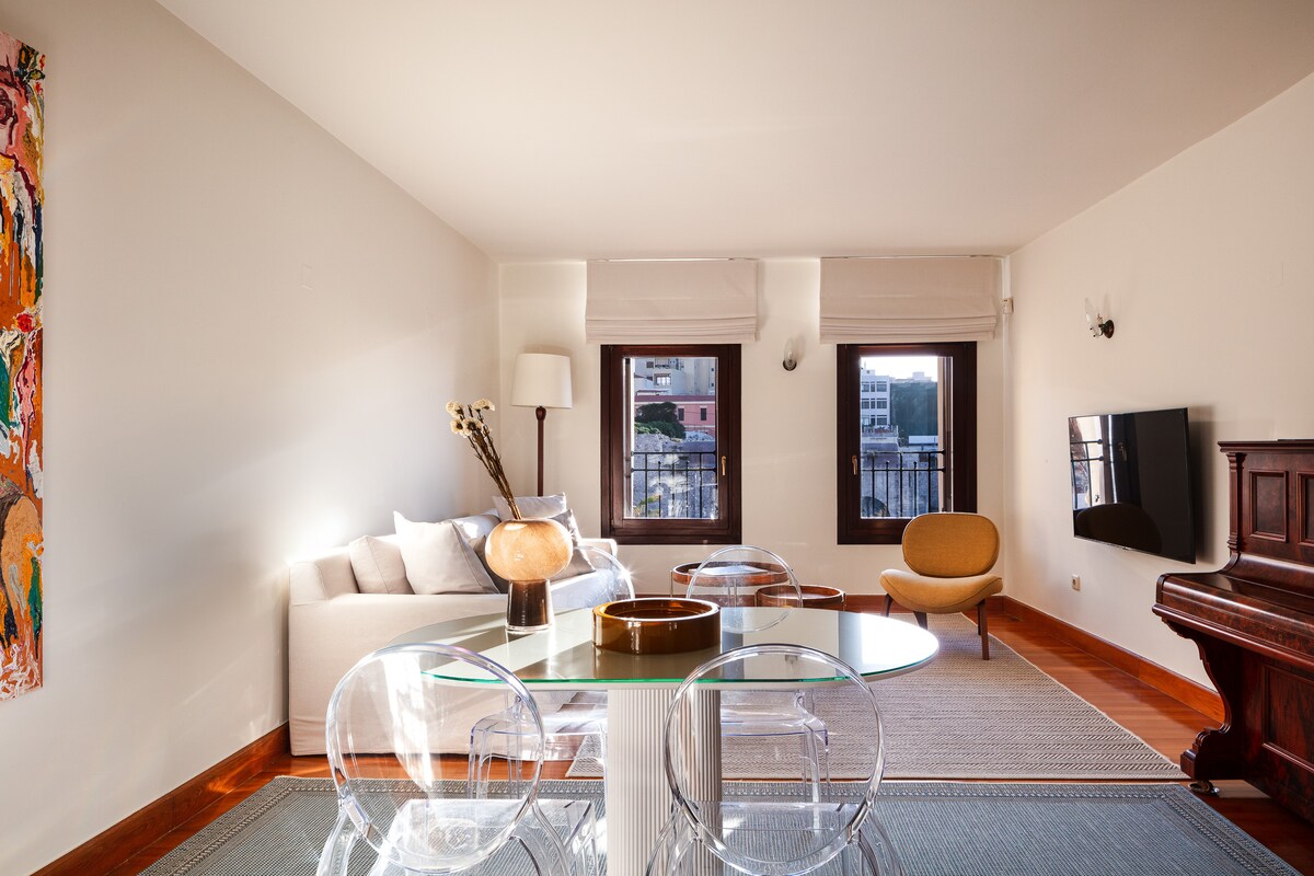 The open living area features high beamed ceilings, large windows, and a light-colored sofa. A round glass dining table accommodates several guests. A piano and an orange accent chair add visual interest. Natural light fills the space, enhancing the overall design.