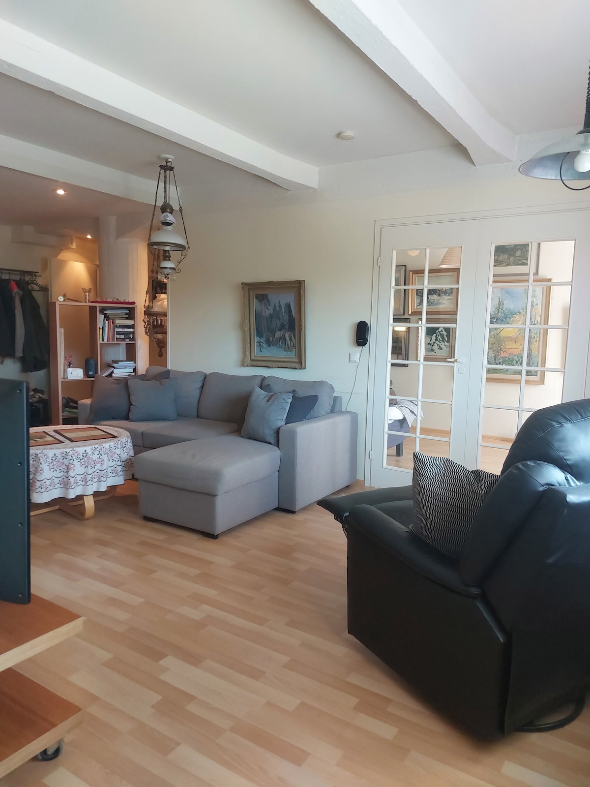 A living area features a comfortable gray sofa and an armchair, providing a relaxed seating arrangement. A small coffee table is centered in the space, with natural light filtering through glass doors that lead to another room. Walls are adorned with framed artwork.