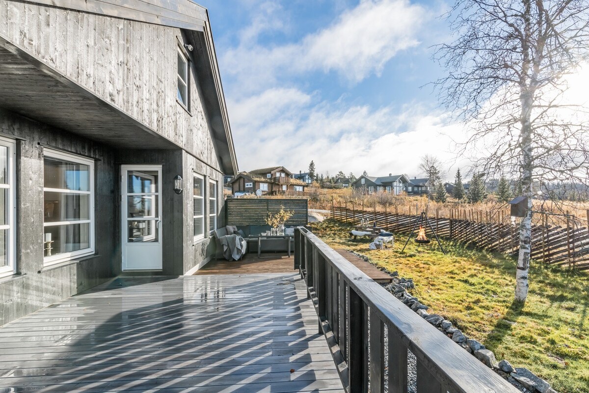 An outdoor deck is presented with wooden planks, leading to an open area featuring a lawn and a small tree. The surrounding landscape includes distant buildings and a low fence, all under a bright sky with patchy clouds.