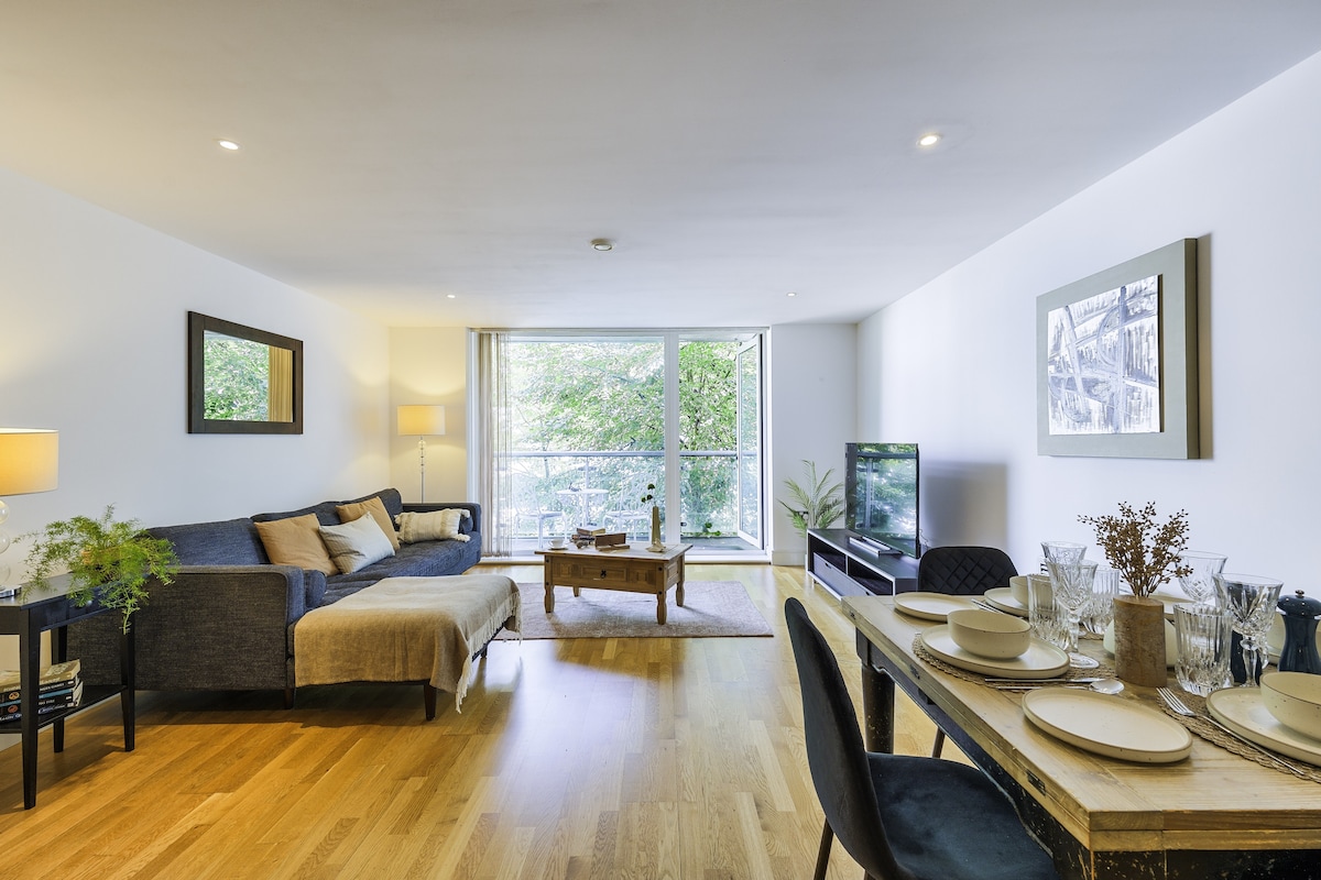 The living area features a spacious arrangement with a comfortable sofa, a wooden coffee table, and a dining table set for four. Large windows invite natural light and offer views of greenery outside. A flat-screen TV is positioned for easy viewing.