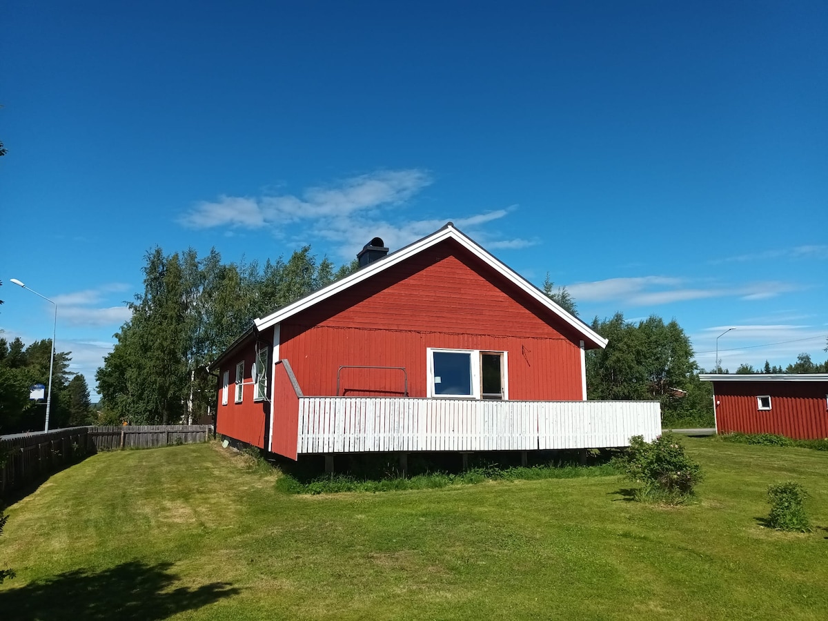 A charming red house is situated on a green lawn under a clear blue sky. The white-washed deck extends from one side of the structure, providing an outdoor space. Surrounding trees and shrubs add touches of nature to the scenic landscape.