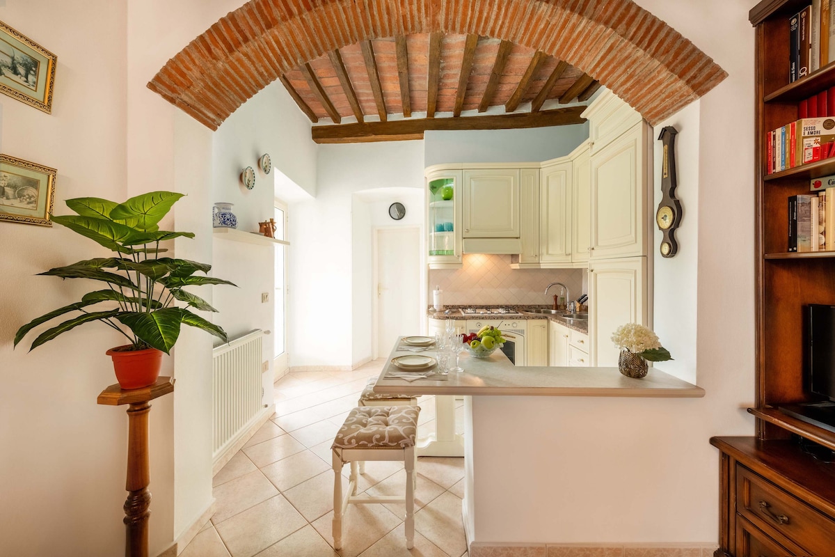 A bright kitchen area is visible, featuring light-colored cabinetry and a marble countertop. A small dining setup is arranged with plates and utensils, while a potted plant adds a touch of greenery. The brick archway and wooden ceiling beams create a warm architectural detail.