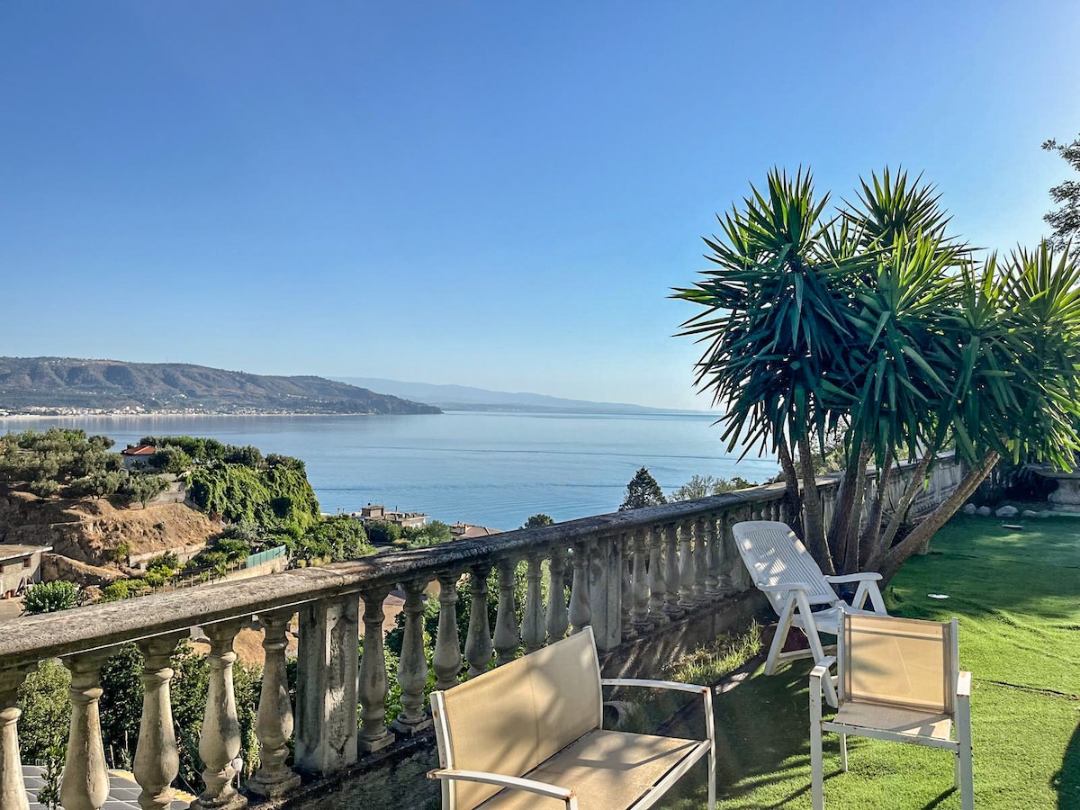 An outdoor terrace features comfortable seating arrangements, including two chairs, overlooking a serene view of the calm sea and distant hills. Lush greenery, including a palm-like plant, adds to the natural ambiance, with clear skies enhancing the panoramic vista.