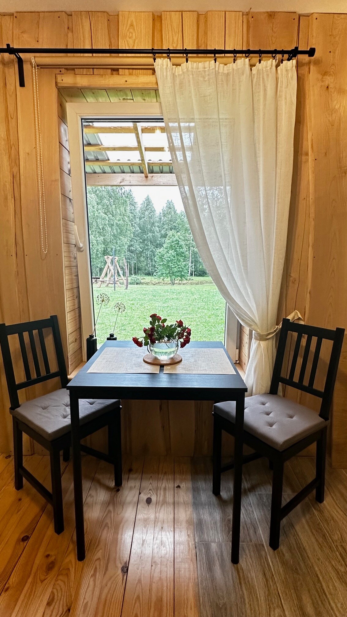 A small dining area is positioned by a window, featuring a black table and two matching chairs. A potted plant decorates the table. A white curtain softly drapes beside the open window, allowing natural light and a view of the greenery outside.