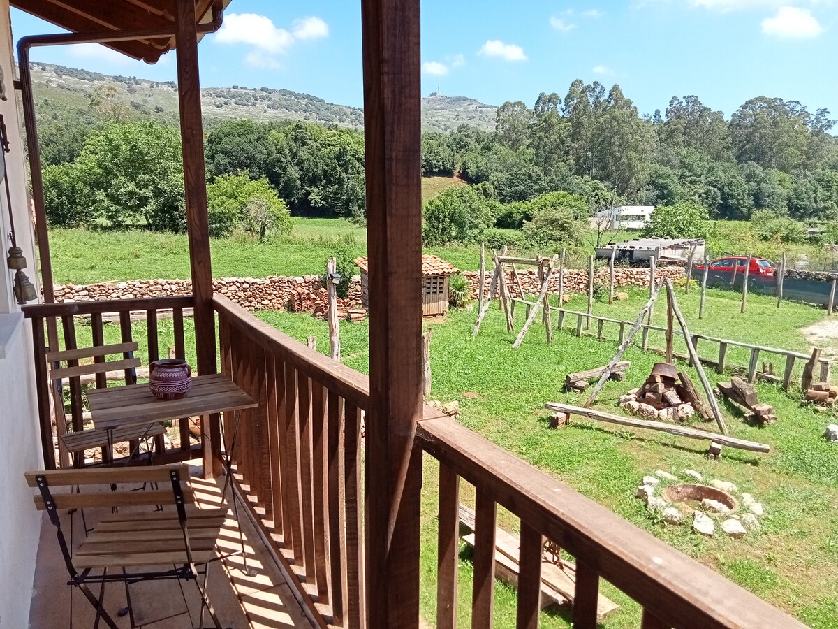 The view from the balcony captures a sprawling green landscape featuring various trees and hillside. A rustic fire pit is visible in the yard, along with wooden structures that add character to the outdoor space. A table and chairs are positioned on the balcony, inviting relaxation.