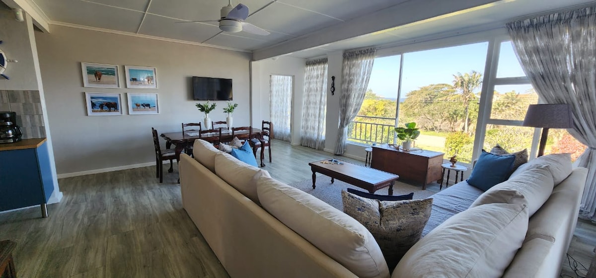 An open-plan lounge and dining area features large windows offering ocean views, allowing natural light to fill the space. A comfortable sofa invites relaxation, while a dining table accommodates several chairs. Framed beach photographs adorn the walls, enhancing the coastal atmosphere.