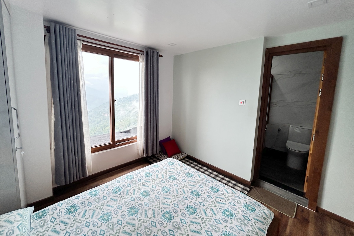 A spacious room features a large bed with a patterned duvet cover and a window providing scenic mountain views. Light-filtering curtains frame the window, while a small seating area is positioned nearby. The bathroom door is visible in the corner, with neutral-toned walls and flooring.