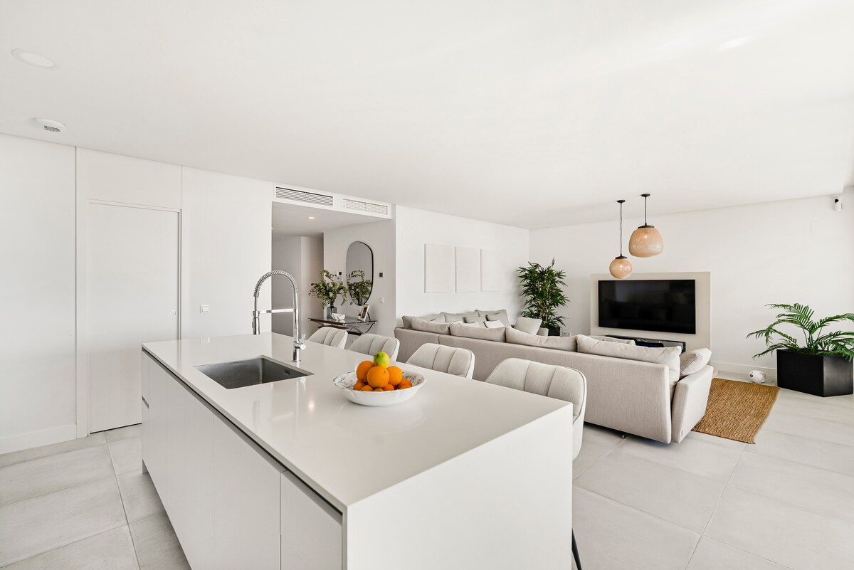 A modern open-plan living area features a sleek kitchen island with a fruit bowl. A light-filled space is created with soft seating arranged around a television, complemented by greenery from potted plants. The neutral-toned decor enhances an inviting ambiance.