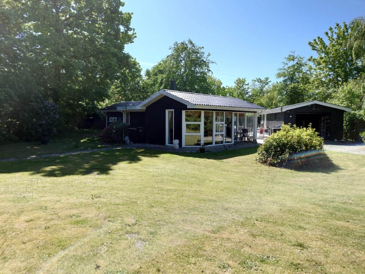 A charming summer house is surrounded by greenery, showcasing a well-maintained lawn. The house features large glass windows that reflect natural light. A separate structure can be seen in the background, adding to the spacious outdoor environment.