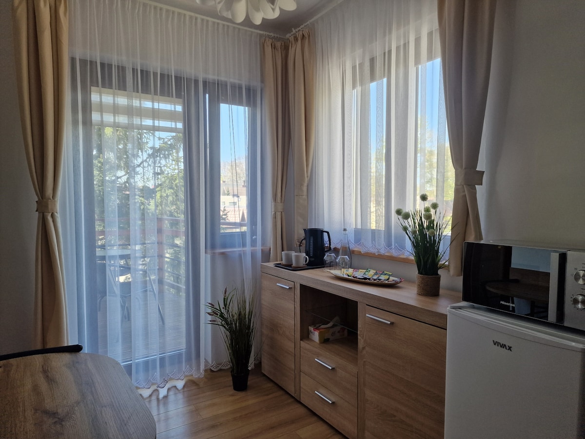 A bright and airy space features large windows adorned with light curtains that allow natural light to fill the room. A wooden cabinet holds a kettle and other items, while a small arrangement of plants adds a touch of greenery. A refrigerator and microwave are also visible.