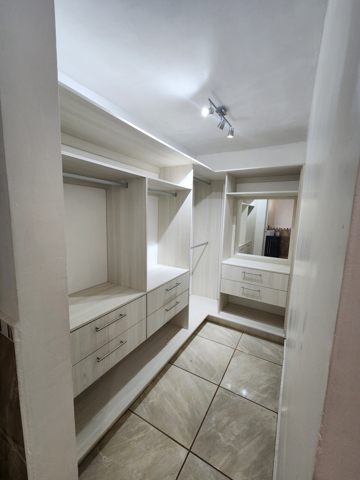 A bright and modern dressing area features built-in cabinetry and mirrors. The space includes drawers and shelves on either side, providing ample storage. The tiled floor reflects light from the overhead fixtures, creating a clean and organized environment.