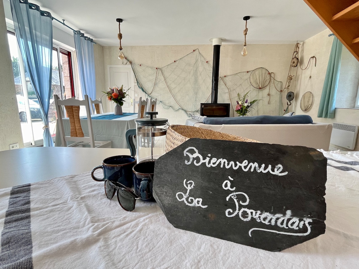A welcoming living space features a slate sign with 'Bienvenue à La Pouardais' prominently displayed on a table. Soft blue curtains frame windows, and a cozy seating area is arranged nearby. Decorative elements, including fishing nets and flowers, enhance the room's charm.