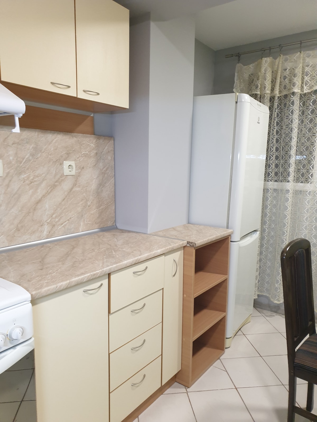 A functional kitchen area is highlighted by light-colored cabinetry and a marble countertop. A refrigerator stands beside the storage shelf, and a stove is positioned nearby. Natural light enters through a window adorned with lace curtains, enhancing the space's practicality.