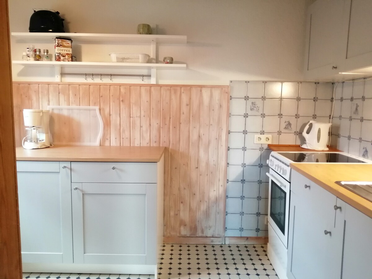 A modern kitchen is depicted, featuring light grey cabinetry and a wood-paneled accent wall. Essential appliances, including a white stove and a kettle, are visible. The patterned flooring adds visual interest, complementing the organized layout with shelves for storage.